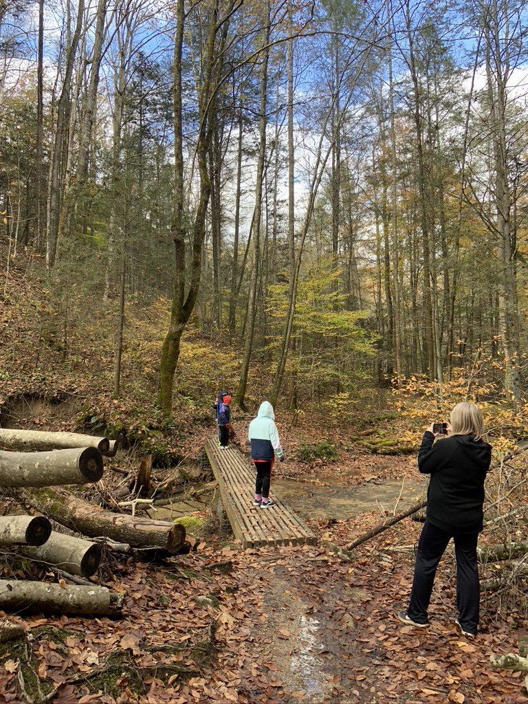 Hikers on a wooden bridge in Red River Gorge, KY. Fall foliage surrounds a mystery hike experience.