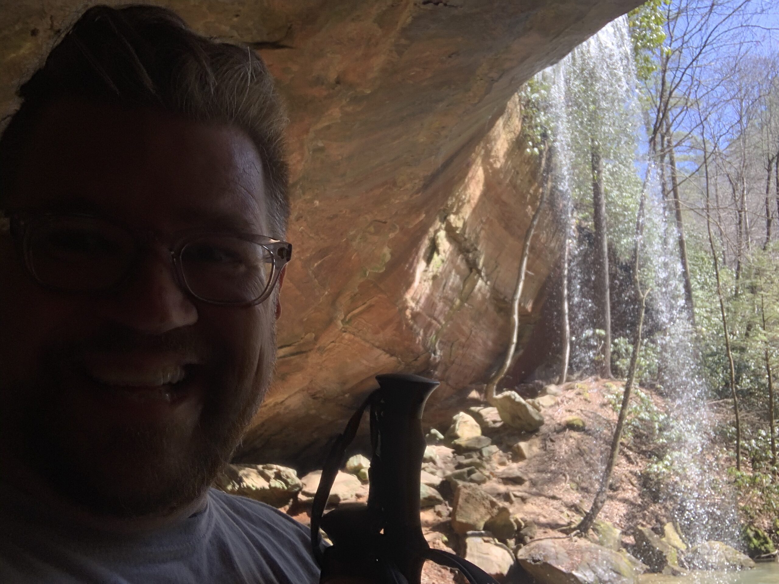 daniel boone national forest red river gorge ky Man smiling under a rock shelter with a waterfall in the Red River Gorge, KY. Hiking stick visible.